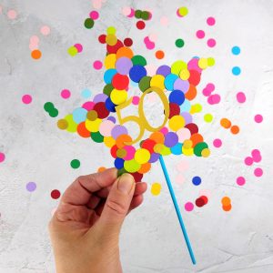 Star Cake Topper is held in model's hand above a table top covered in rainbow dot paper confetti. Star is covered in rainbow coloured paper circles and has number 50 in gold italic script in the centre.