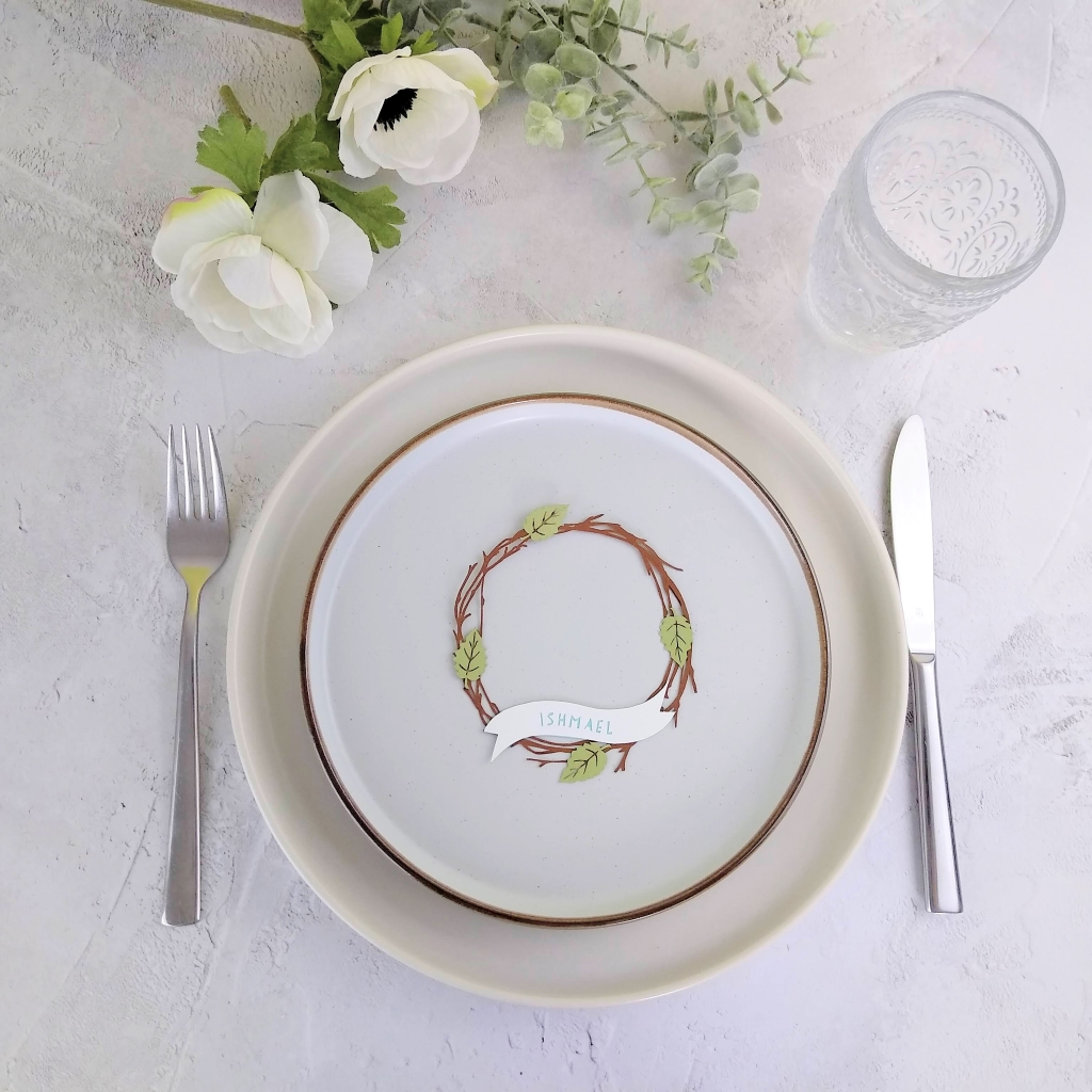 Personalised Papercut wreathplace setting shown on a grey plate as part of formal table setting. Name Ishmael is cut out of pale blue letters on personalised banner across the wreath.