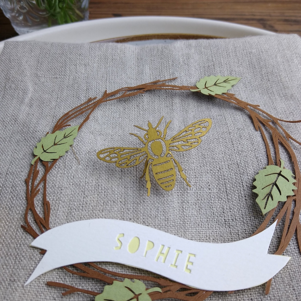 Papercut wreath of brown birch twigs with 4 light green leaves. At the base of the wreath is a white paper banner with the name Sophie cut out in lemon yellow letters. In the centre of the wreath is a gold honey bee confetti piece raised to make it look as though it is taking flight.
