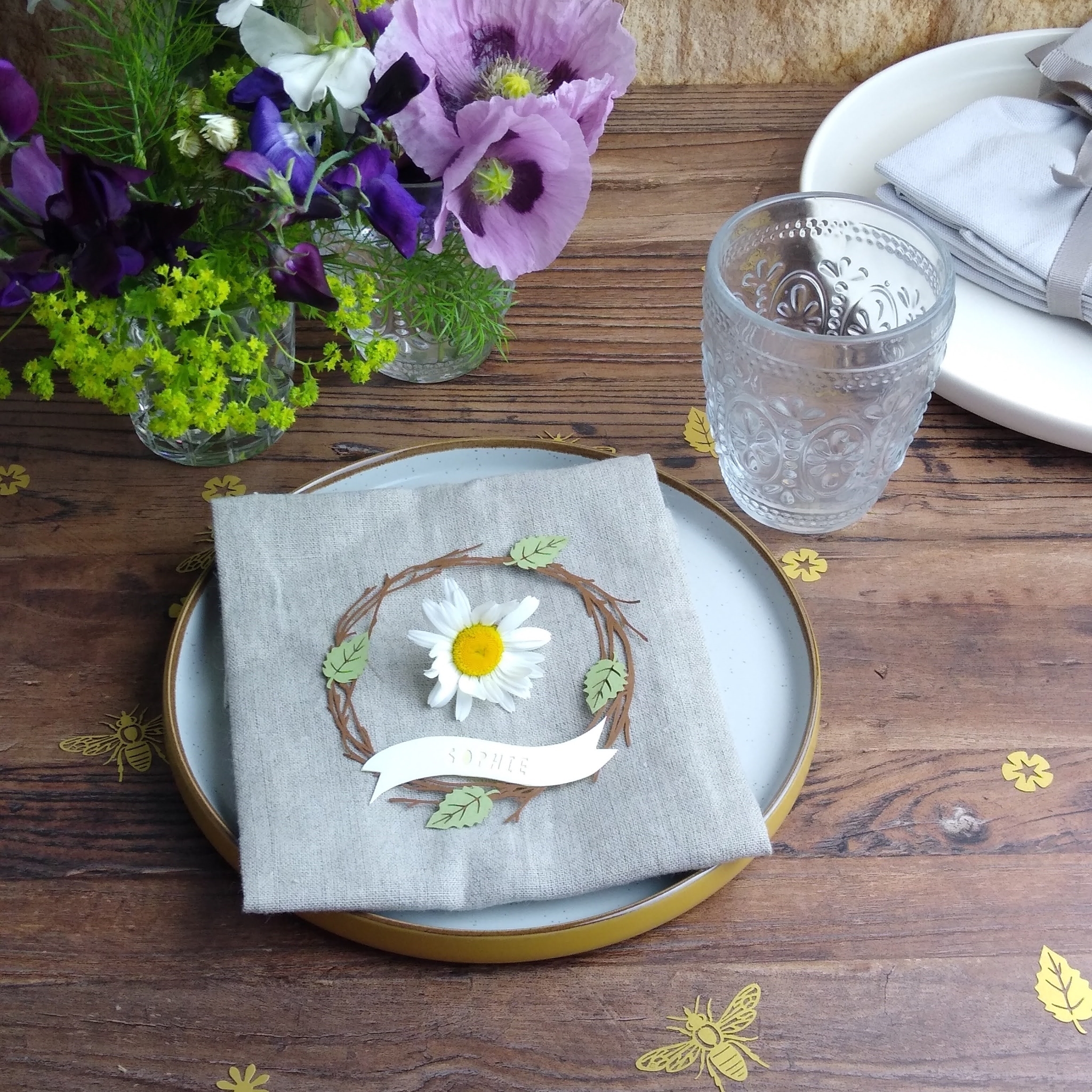 Papercut birch wreath place setting placed on linen napkin with a white daisy placed in the centre. Place setting is on a plate and the table set up for a special occasion with fresh flower arrangement and formal tableware.