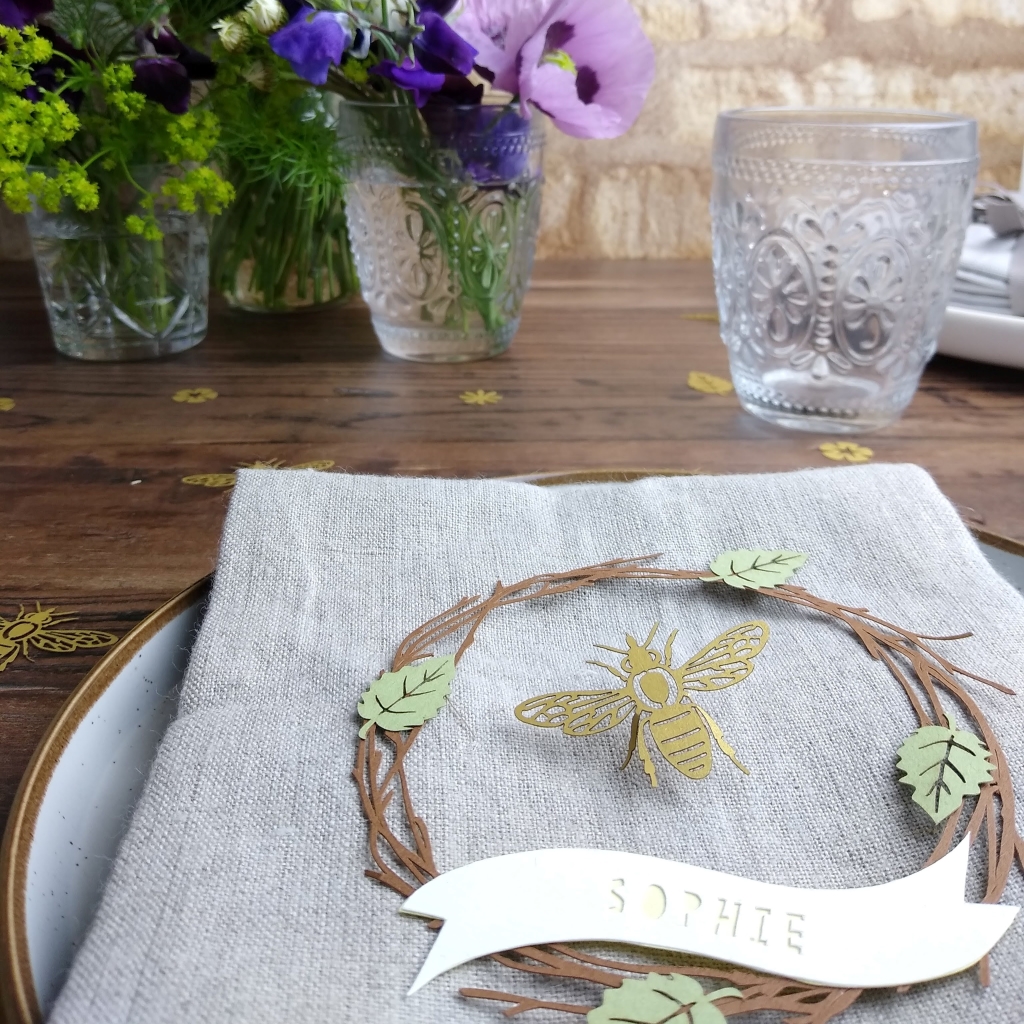Papercut wreath place setting with the personalisation Sophie in yellow letters. Placed on napkin with honey bee confetti piece in centre of place setting.