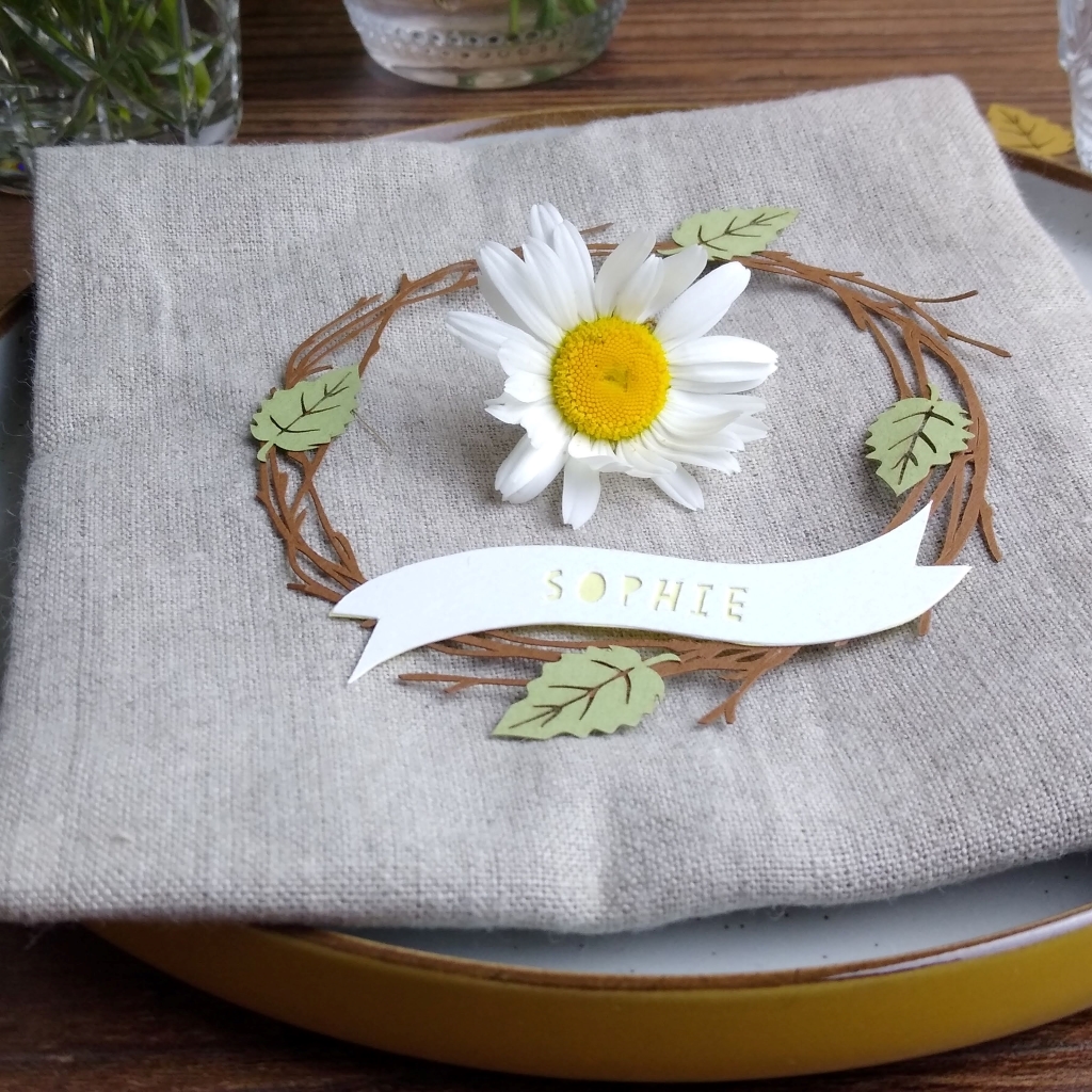 Papercut birch twig wreath with 4 light green leaves (at positions north, south, east and west on wreath), sits on a beige linen napkin. A white daisy is placed inside the papercut wreath. Personalisation reads Sophie in yellow letters.