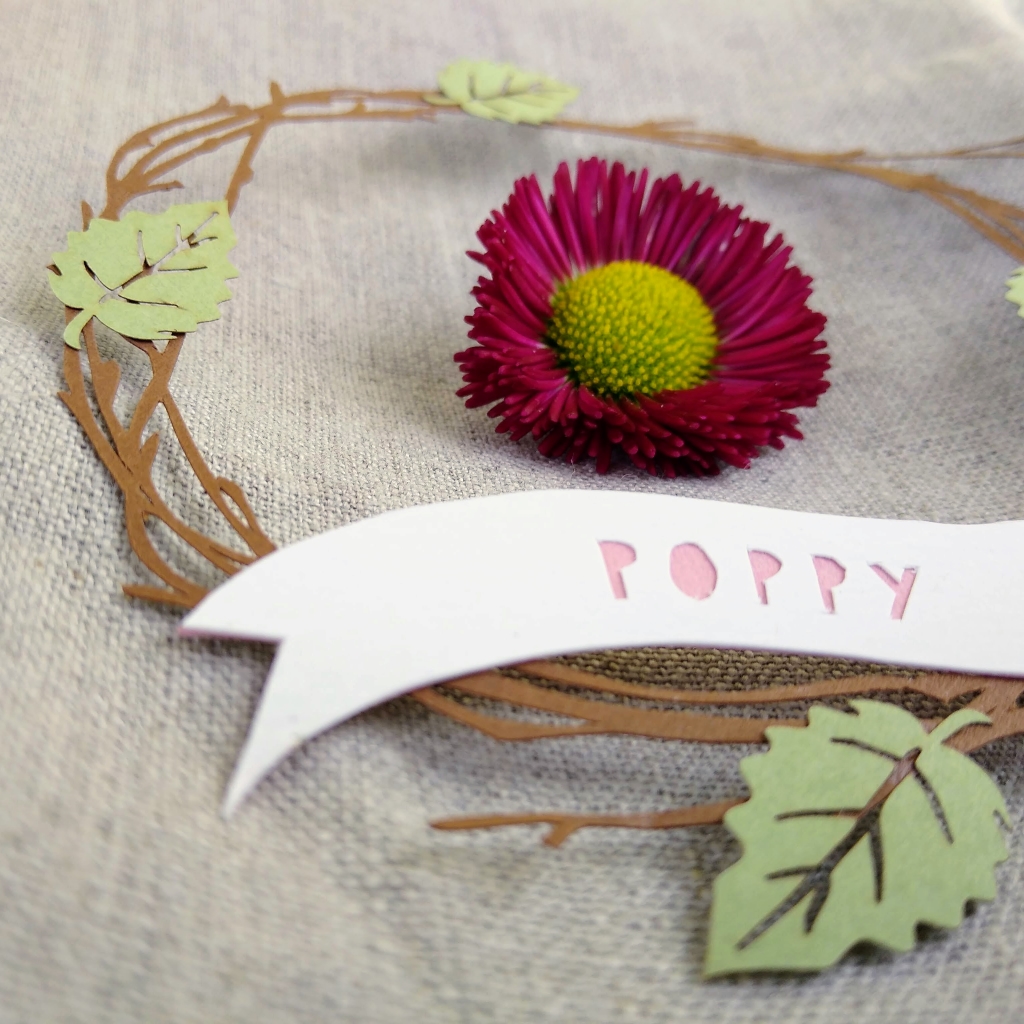 Papercut wreath of brown birch twigs with 4 light green leaves. At the base of the wreath is a white paper banner with the name Poppy cut out in dusky pink letters.