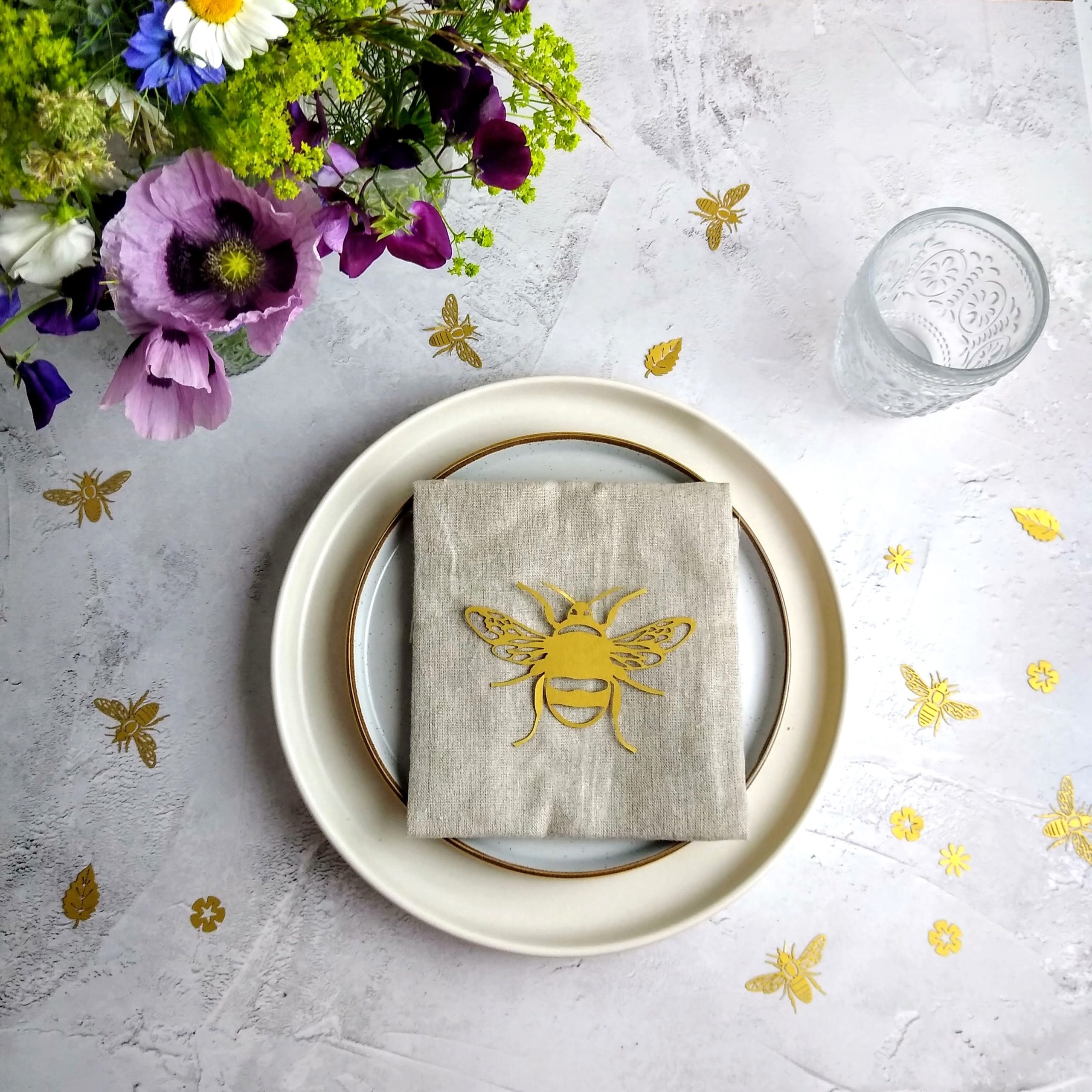 Gold Paper Bumble Bee Place Setting on linen napkin and cream tableware. Table is decorated with fresh flowers and gold bee and flower table confetti