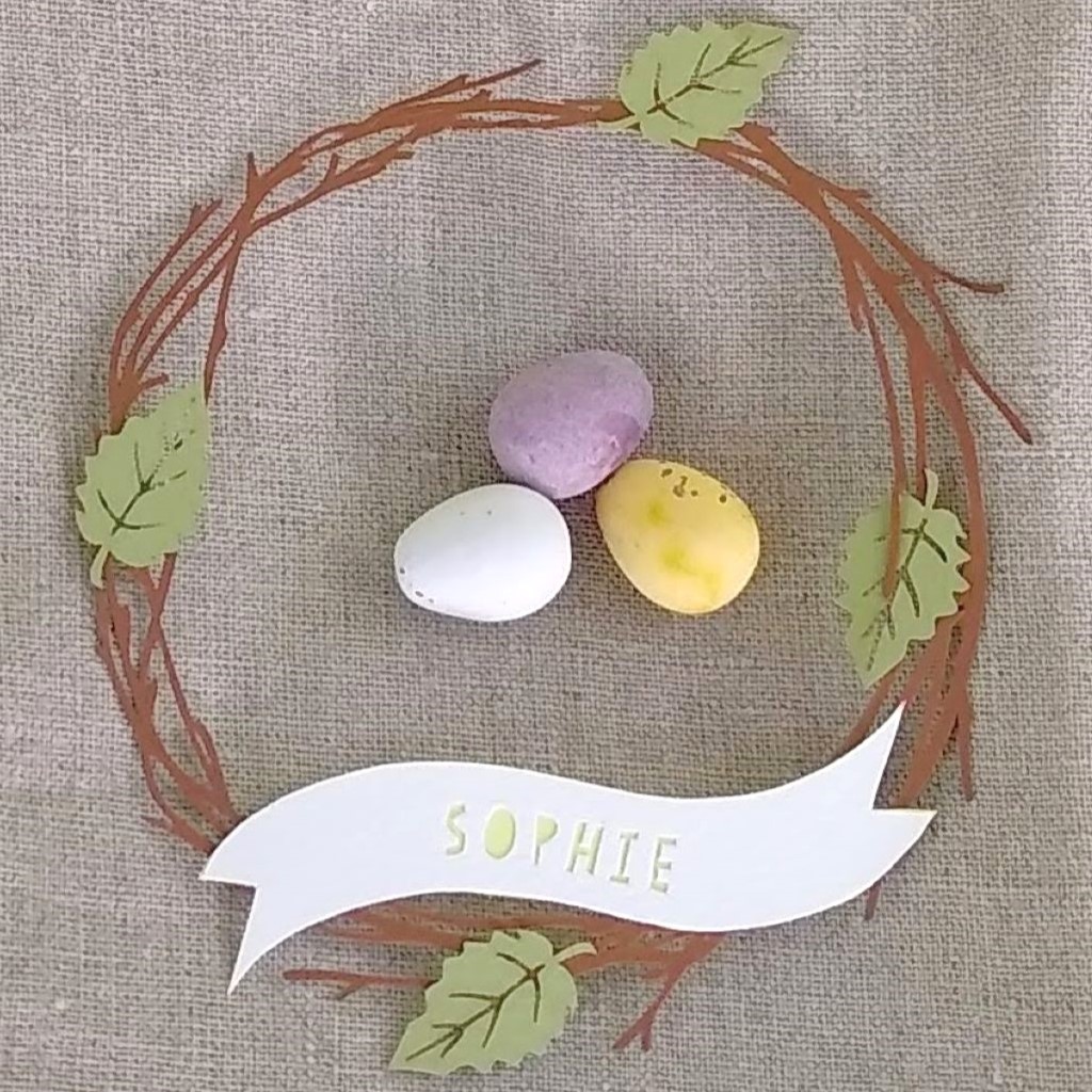 Closeup of papercut brown birch twig wreath with 4 light green leaves. White paper banner across bottom of wreath reads Sophie cut in pale yellow letters.