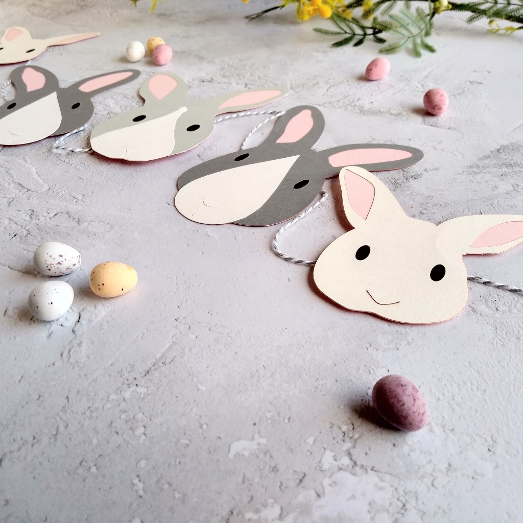 A closeup image of one white bunny face decorations, one dark grey and white rabbit face and one pale grey bunny decoration. There is a v shaped line to depict the bunny nose on each decoration.