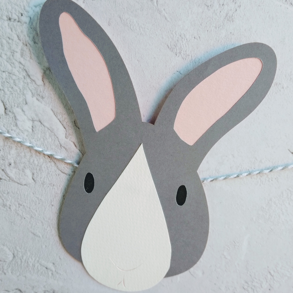 Dark grey bunny face with white tear drop shaped nose patch. Small oval black eyes and pale pink ears