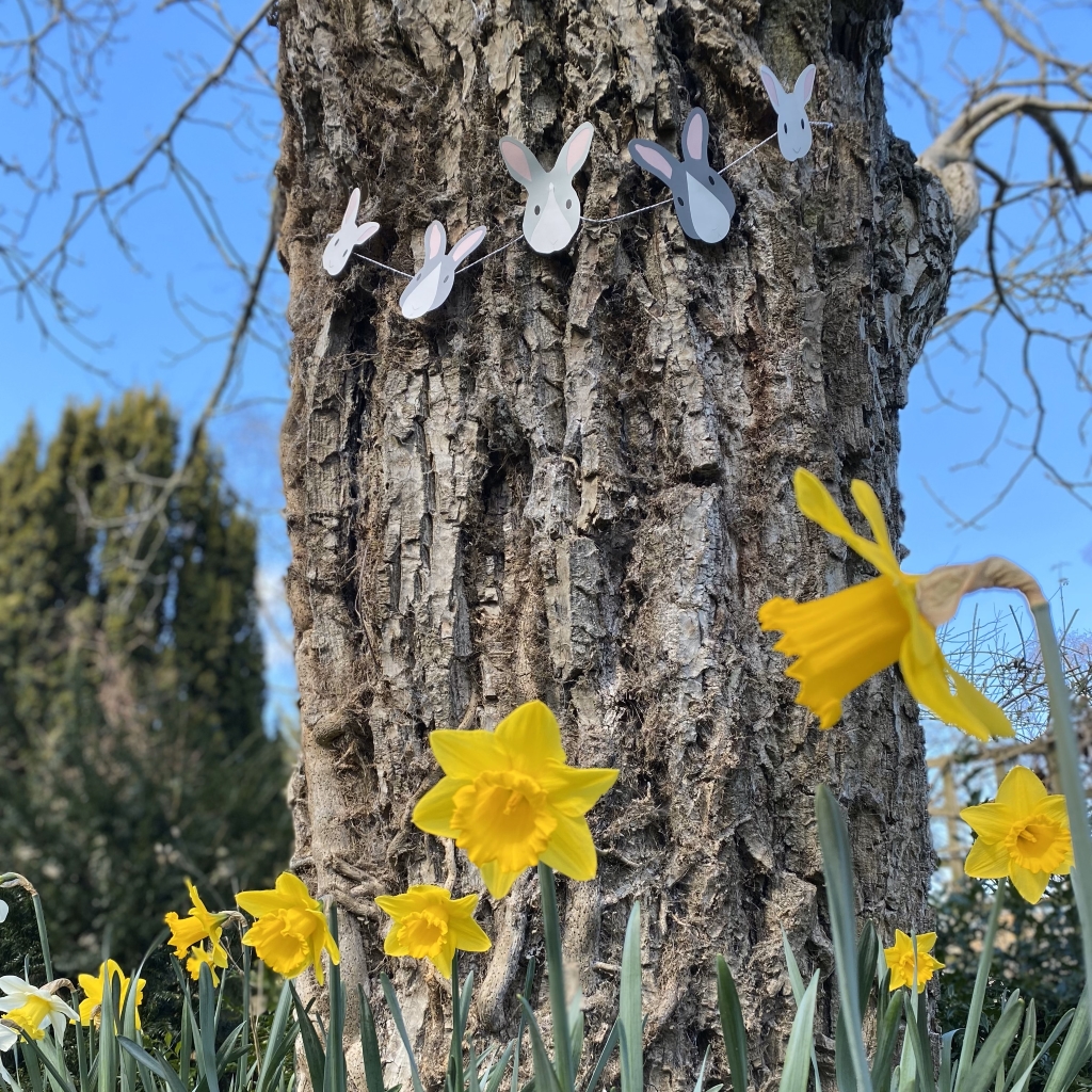 Easter bunny garland is shown wrapped around a large tree trunk in a garden with daffodils at the base of the tree.