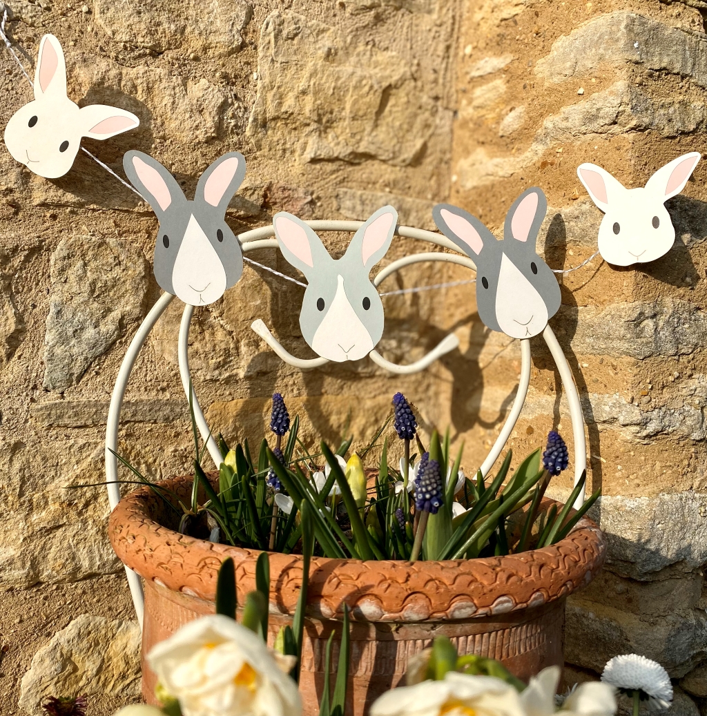 Easter bunny paper garland shown displayed outside against a garden wall above a flower pot filled with spring flowers.