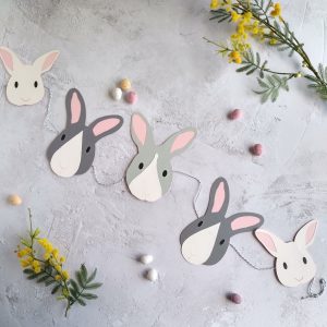 Easter bunny paper garland displayed flat. Garland has 5 bunny face decorations threaded onto grey and white bakers twine which is tied in a loop at each end. At each end of the garland is a white bunny face with black eyes and pale pink ears. Next are dark grey bunnies that have a white nose patch, pale pink ears and black eyes. The central bunny is pale grey with a wihte face strips, black eyes and pale pink ears.