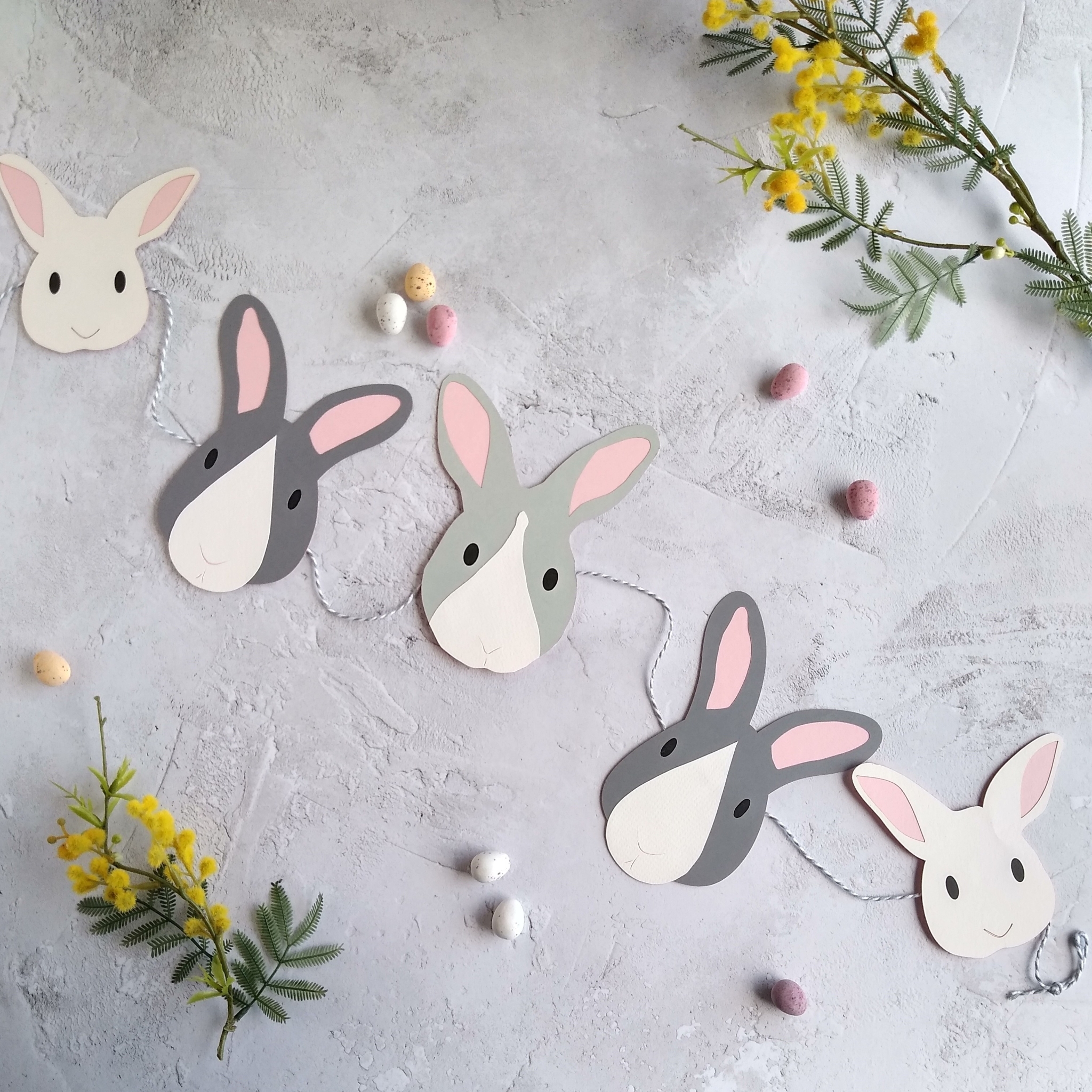 Easter bunny paper garland displayed flat. Garland has 5 bunny face decorations threaded onto grey and white bakers twine which is tied in a loop at each end. At each end of the garland is a white bunny face with black eyes and pale pink ears. Next are dark grey bunnies that have a white nose patch, pale pink ears and black eyes. The central bunny is pale grey with a wihte face strips, black eyes and pale pink ears.