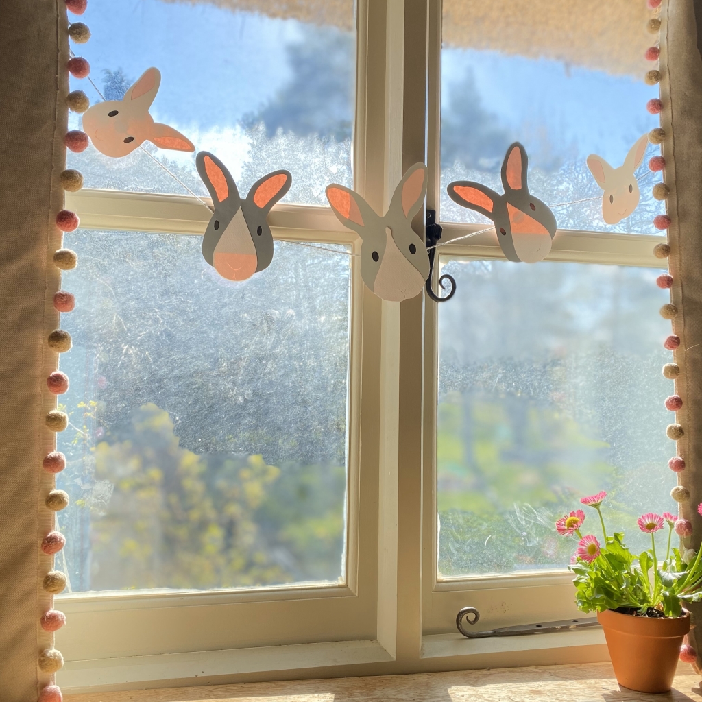 Eastr bunny garland is displayed hanging across a window with white wooden frames. Through the window you can see a garden on a sunny day.