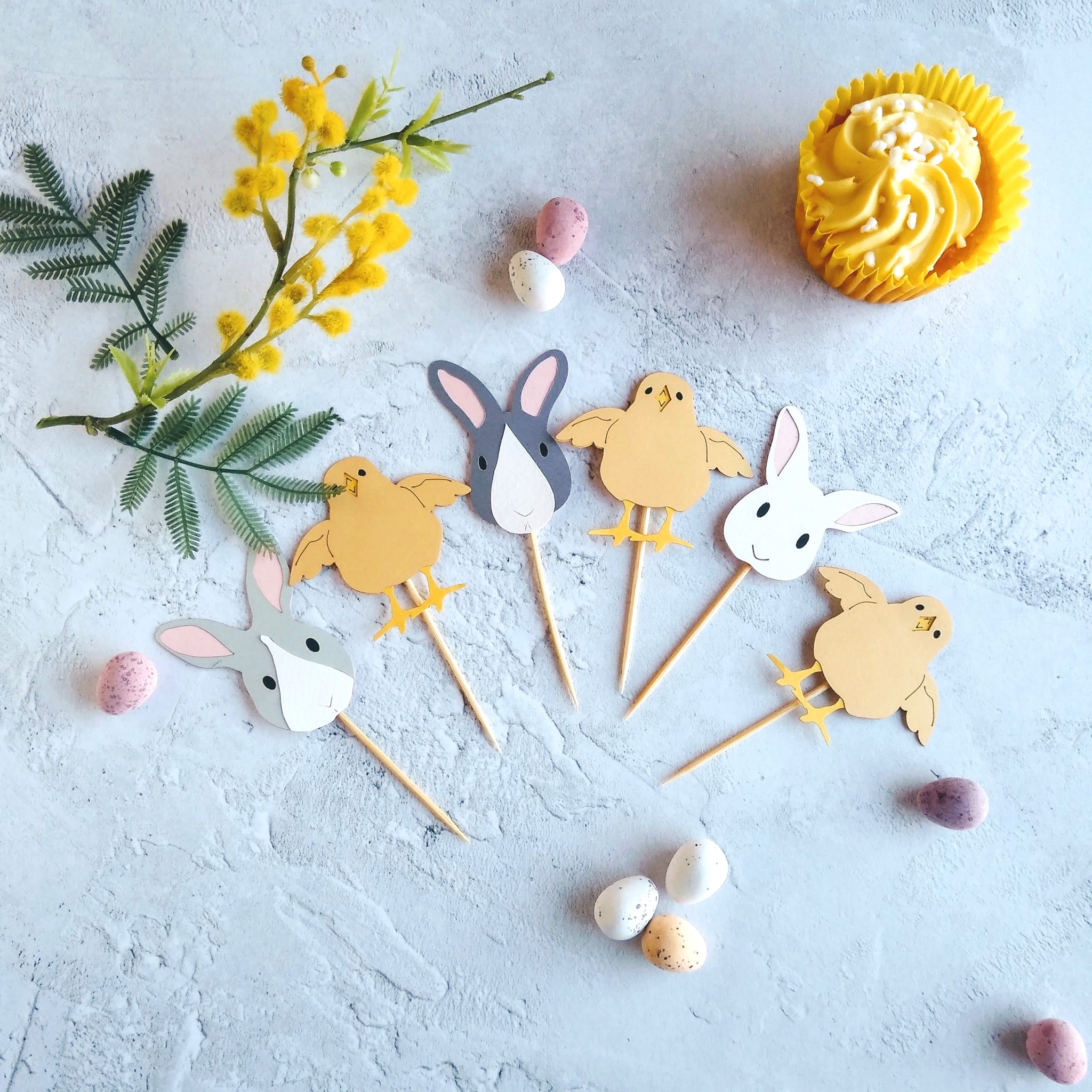 6 Easter themed cupcake toppers spread in an arc. 3 cupcake toppers are identical pale yellow chicks with outstretched wings. 3 cupcake toppers are Easter Bunny faces - one all white with pink ears and black ees, one pale grey and white with pink ears and black eyes, one dark grey and white with pink ears and black eyes.