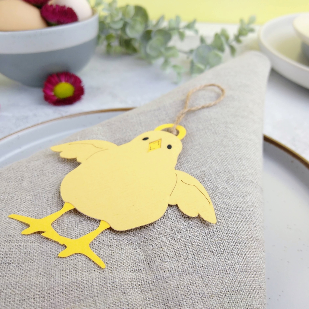 Easter Chick Design 2 displayed on beige linen napkin as a keepsake place setting. Chick is pale yellow with dark yellow legs and beak. Wings are outstretched. Chick is threaded with a 5cm hanging loop of jute twine.