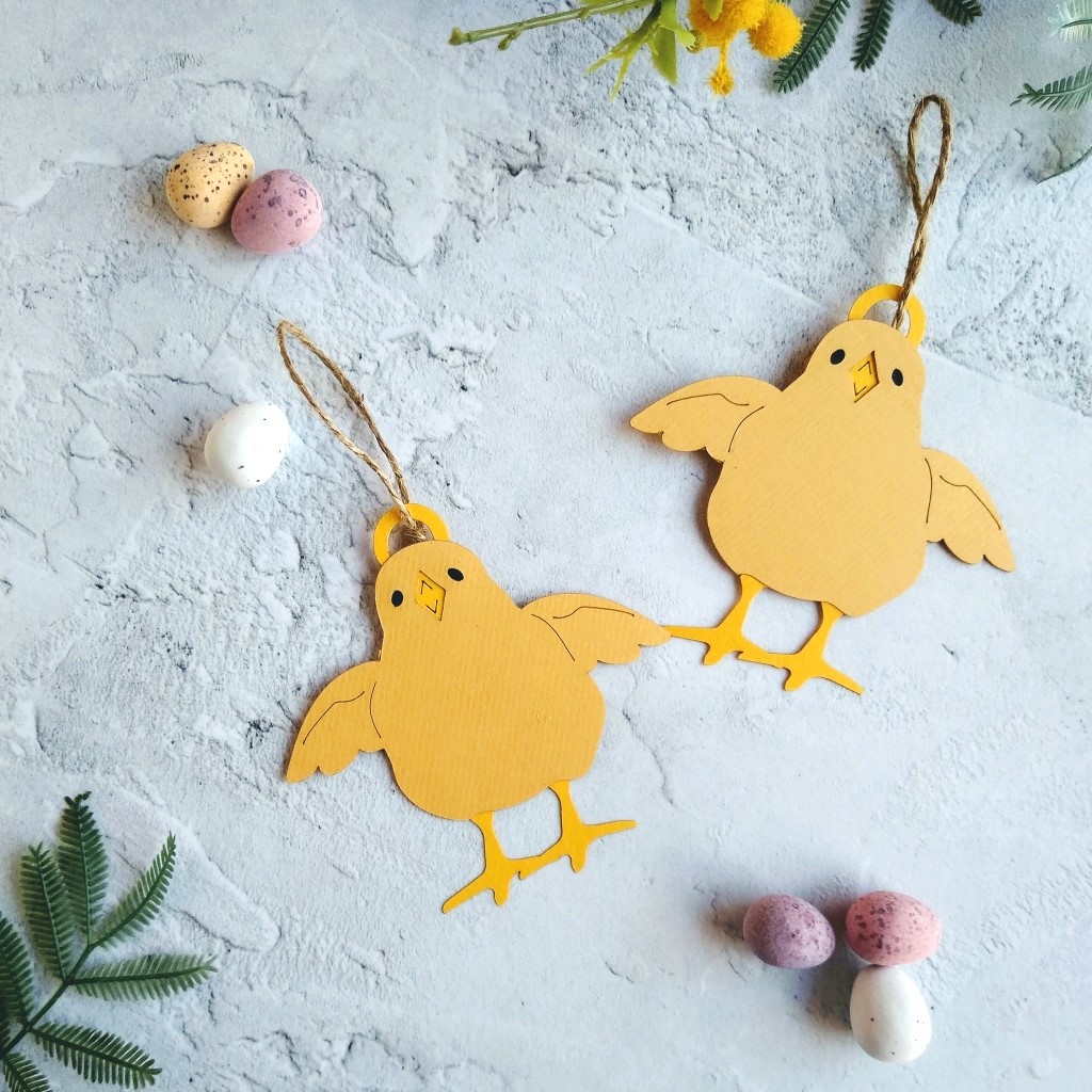 Pair of Easter Chick Hanging Decorations deisgn two laid on table with yellow mimosa flowers and mini eggs. Chicks are pale yellow with dark yellow legs and beak. Wings are outstretched and beak is open. Chick have 5cm jute twine hanging loop.