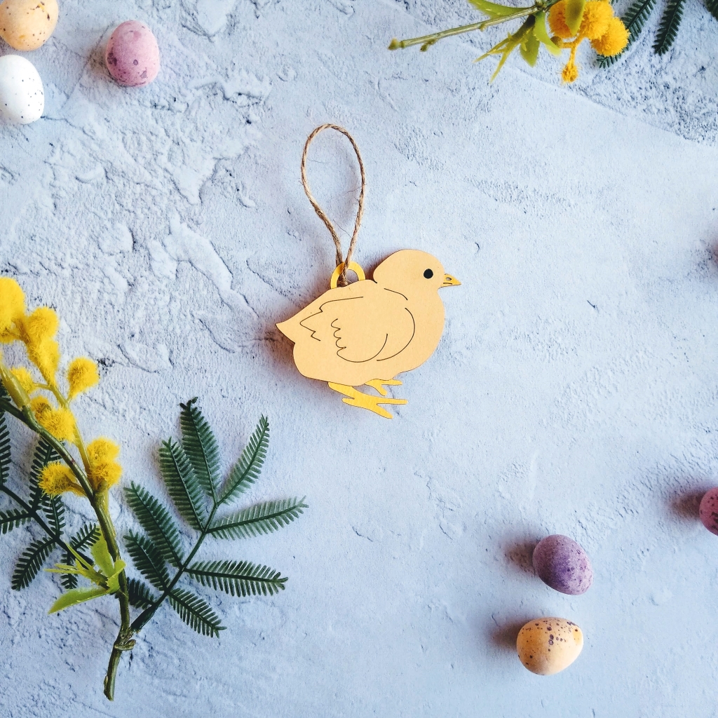 Design One Easter Chick Hanging Decoration. Chick is made from pale yellow paper with dark yellow legs and closed beak. Chick has wing details, a small black eys and is in a crouched position. Decoration is threaded with 5 cm jute twine loop