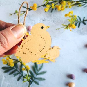 Pair of Easter Chick Hanging decorations surrounded by yellow mimosa flowers. One chick is held in models hand