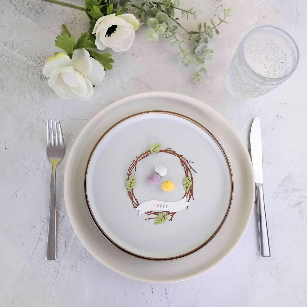 Papercut wreath of twigs and leaves is used as a place setting on a plate. 3 mini eggs are placed inside the wreath place setting. Personalisation reads "poppy" in pink letters.