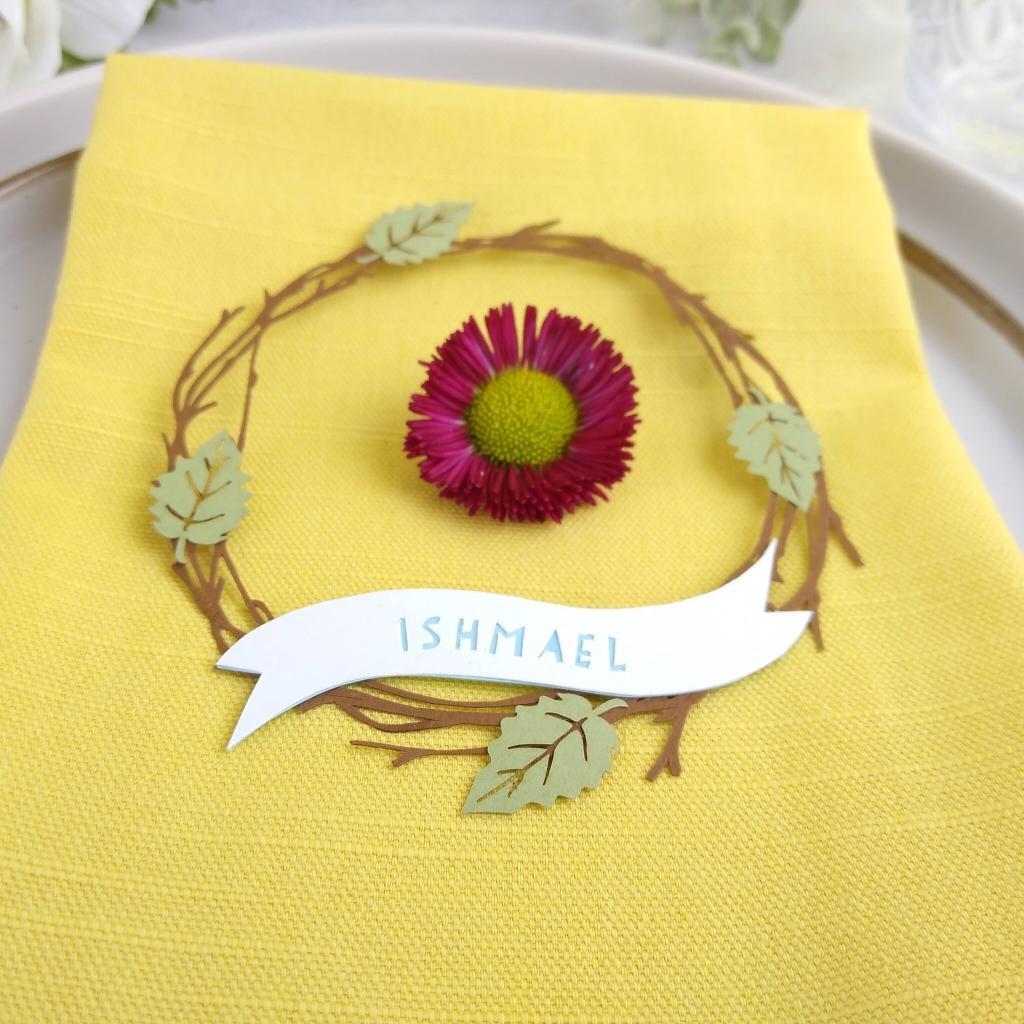 Papercut birch twig wreath with 4 light green leaves (at positions north, south, east and west on wreath), sits on a bright yellow linen napkin. A pink daisy is placed inside the papercut wreath. Personalisation reads Ishmael in blue letters.