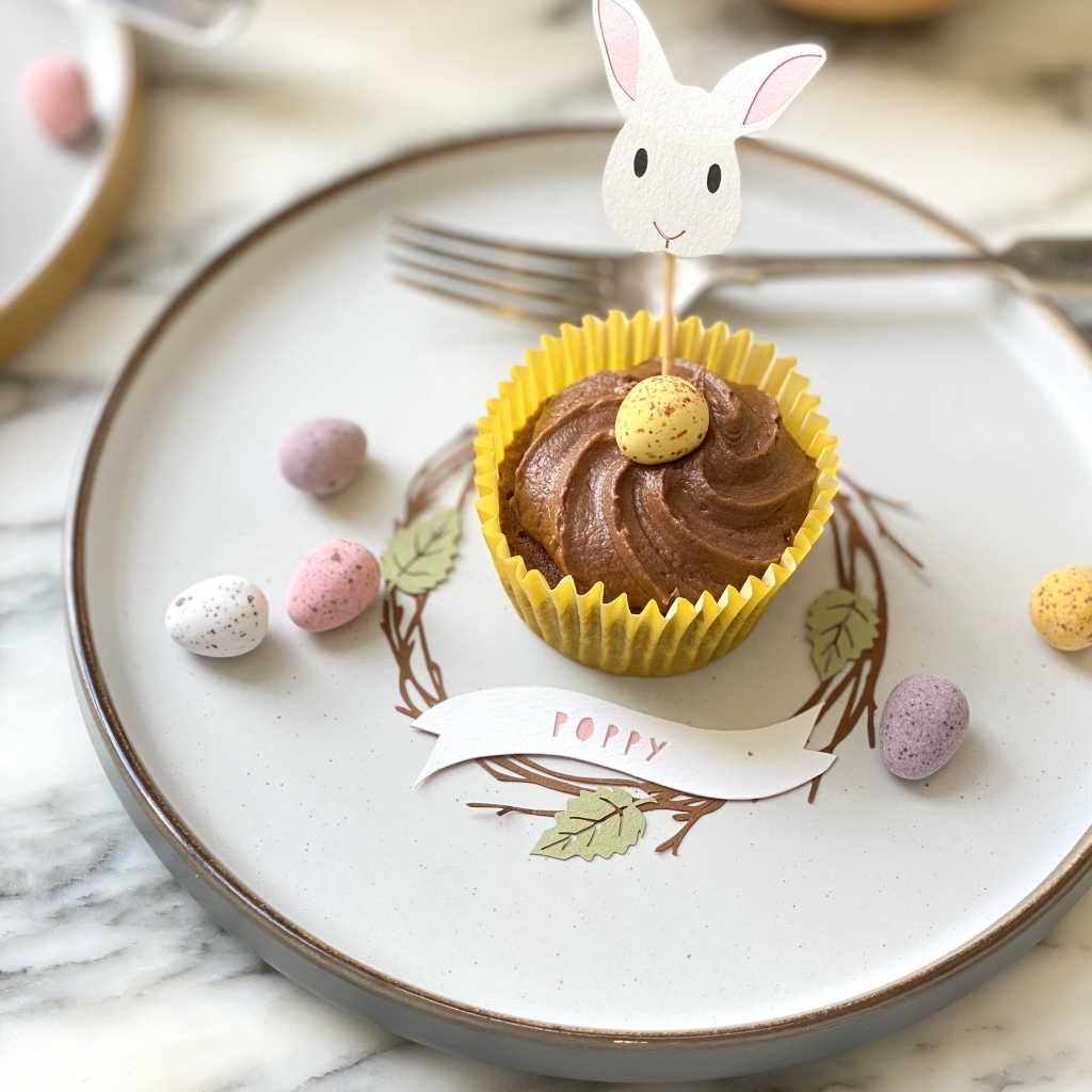 Papercut wreath place setting with the name poppy in pink. Place setting is on a plate decorated with mini eggs. A cupcake with a paper bunny cake topper is placed in the middle of the place setting.