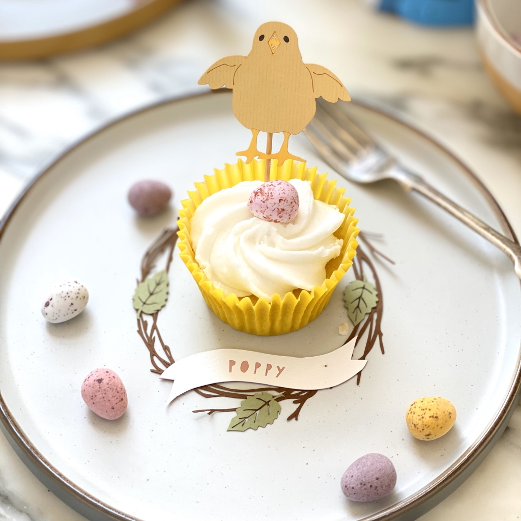 Papercut wreath place setting with the name poppy in pink. Place setting is on a plate decorated with mini eggs. A cupcake with a chick cake topper is placed in the middle of the place setting.