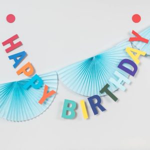 Happy birthday garland strung across a white wall which is also decorated with a blue fan garland. Happy birthday garland letters are all different colours including shdes of red, orange, blue, yellow and green. The garland has a bright red circle at each end.