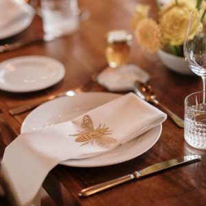 Gold Honey Bee Place setting on top of a white napkin. Napkin is placed in the centre of a white plate on a dark wood table with cutlery, glassware, flowers and a jar of honey as a wedding favour.