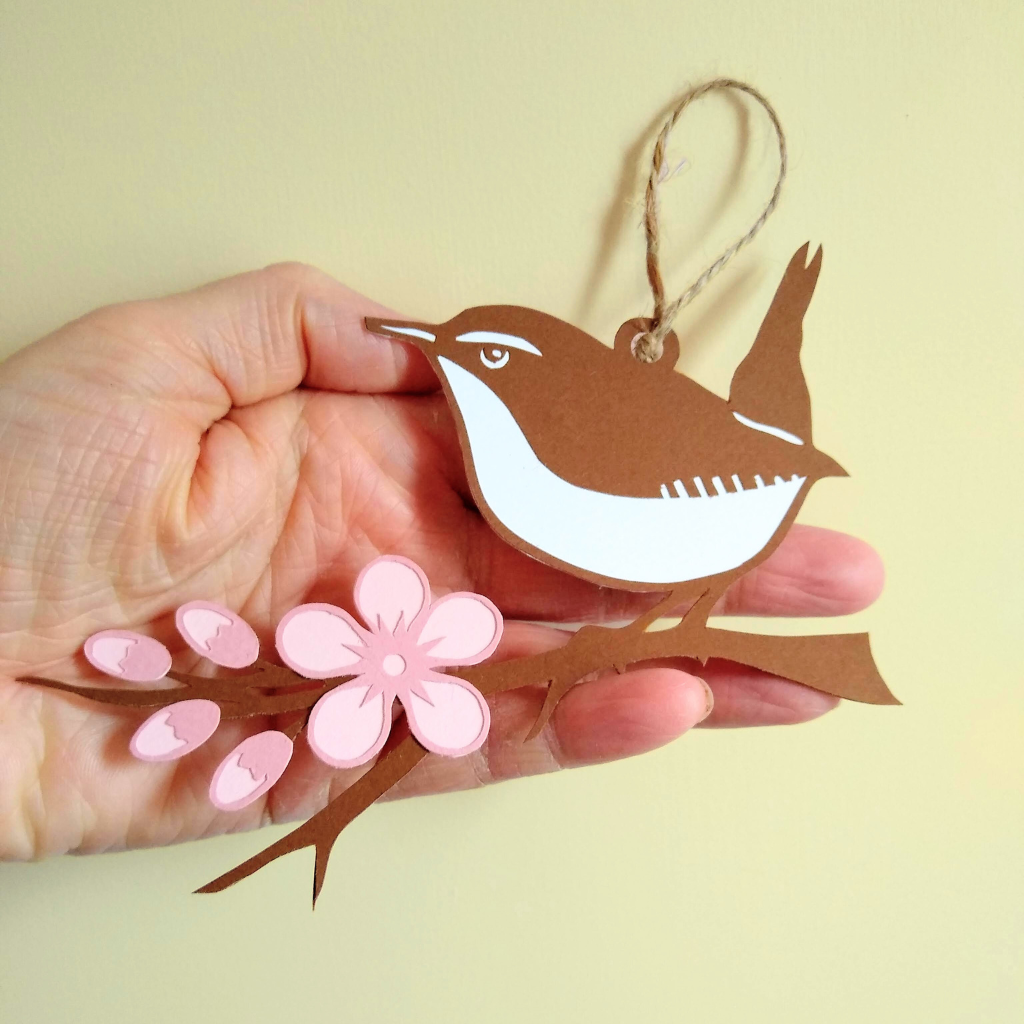 Large size papercut wren hanging decoration held in model's hand. Decoration reaches from tip of model's middle finger to the end of their palm