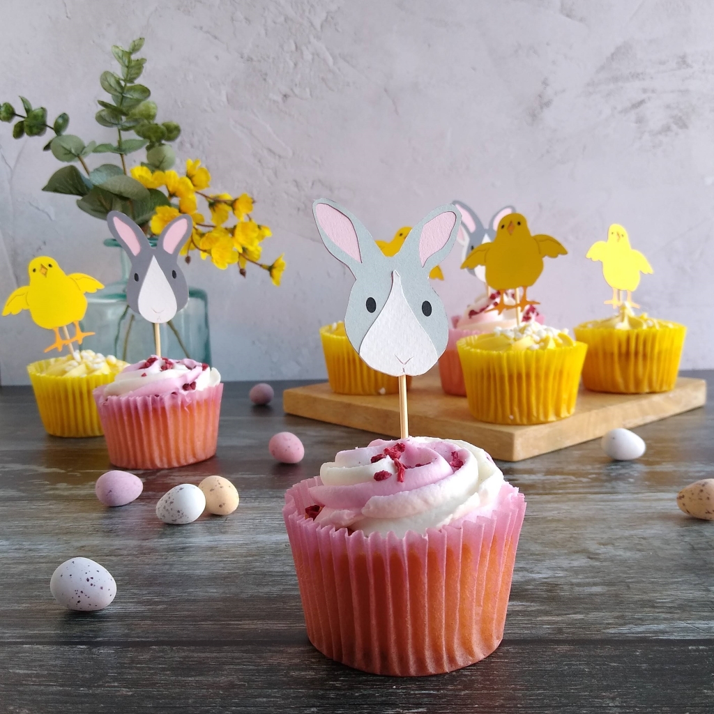 Pale grey and white Easter bunny cupcake topper displayed in pink cupcake infront of a selection of cupcakes with bunny or chick cupcake toppers.