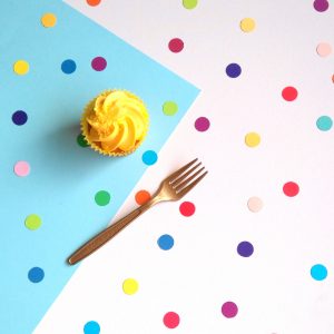 Overhead view of rainbow dot confetti spread evenly across a party table decorated with white and blue tablecloth. Iced cupcake and gold fork are also on the party table.