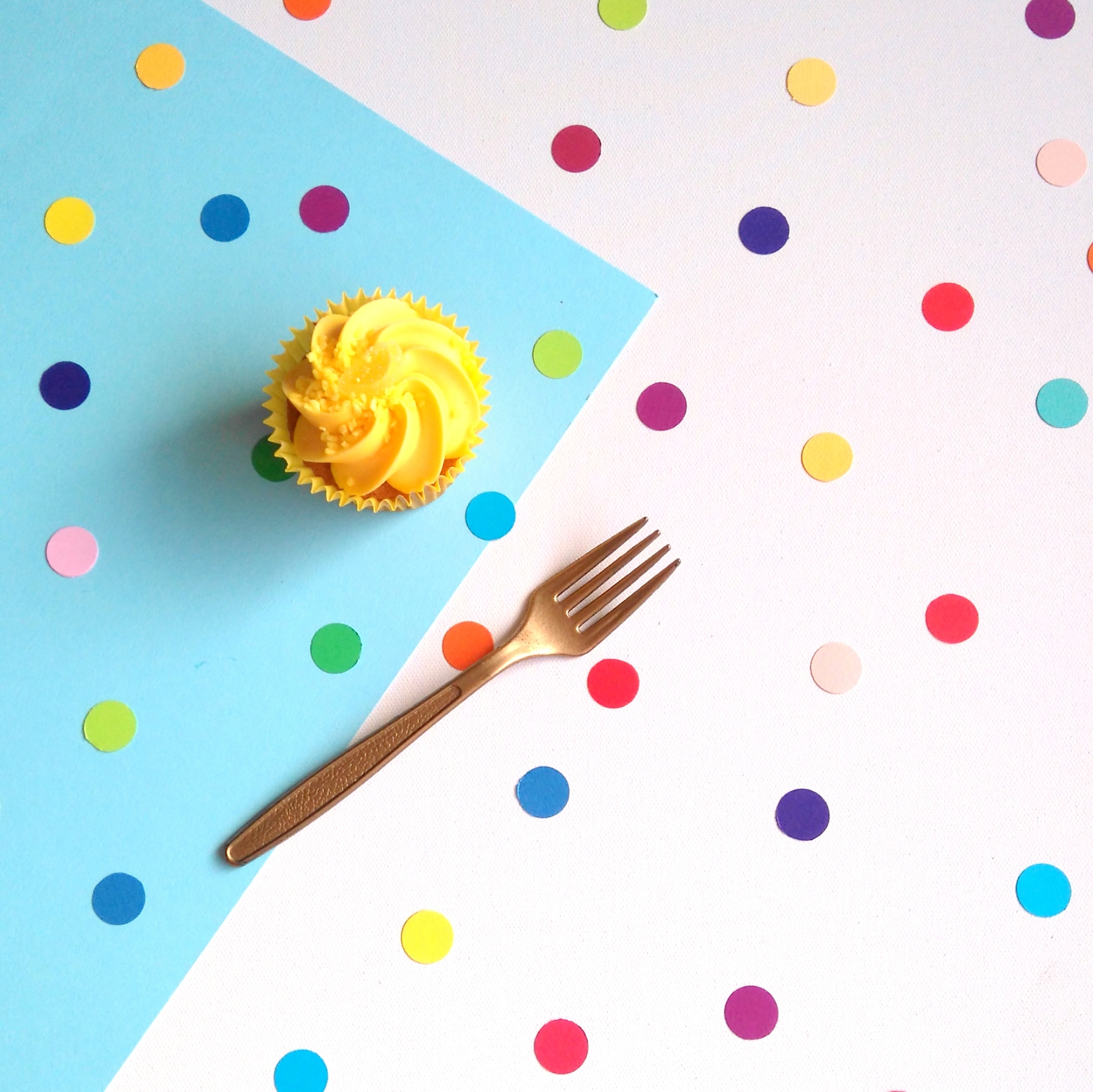 Overhead view of rainbow dot confetti spread evenly across a party table decorated with white and blue tablecloth. Iced cupcake and gold fork are also on the party table.