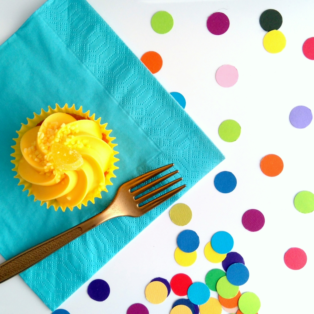 Rainbow dot confetti sprinkled close together on a party table. Iced cupcake and gold fork sit on party table with a blue paper napkin.
