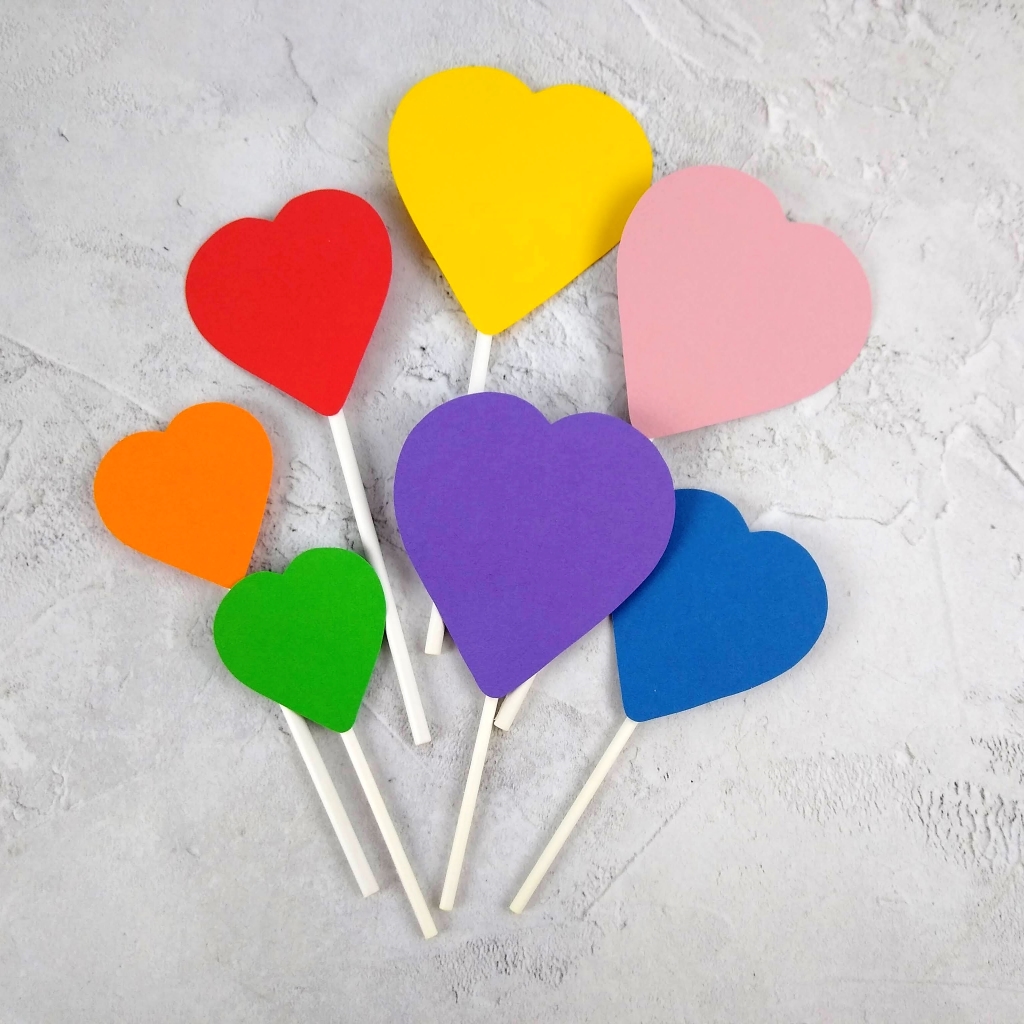 Set of 7 heart shaped cake toppers laid out on a grey table top. Hearts are different sizes, each is attached to a white paper lollipop stick. Heart colours are red, orange, yellow, green blue, purple and pink.