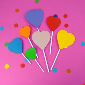 Set of 7 heart shaped cake toppers laid out on a bright pink backbround surrounded by rainbow dot confetti. Hearts are different sizes, each is attached to a white paper lollipop stick. Heart colours are red, orange, yellow, green blue, purple and pink.