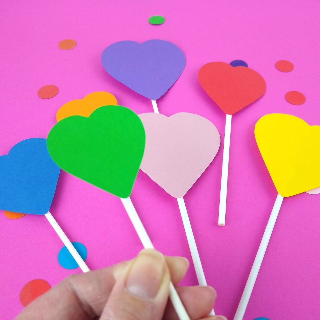 Model holds green heart cake topper in hand above the rest of the cake topper set which is lying on a bright pink background.