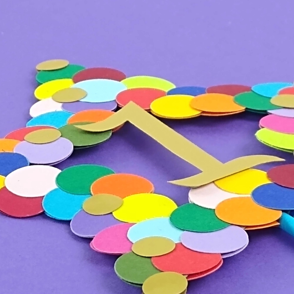 Close view of rainbow star cake topper. Outline of 5 pointed star is covered on both sides in paper circles in all the colours of the rainbow along with smaller gold circles. Number 1 in gold paper is attached to the centre of the star.