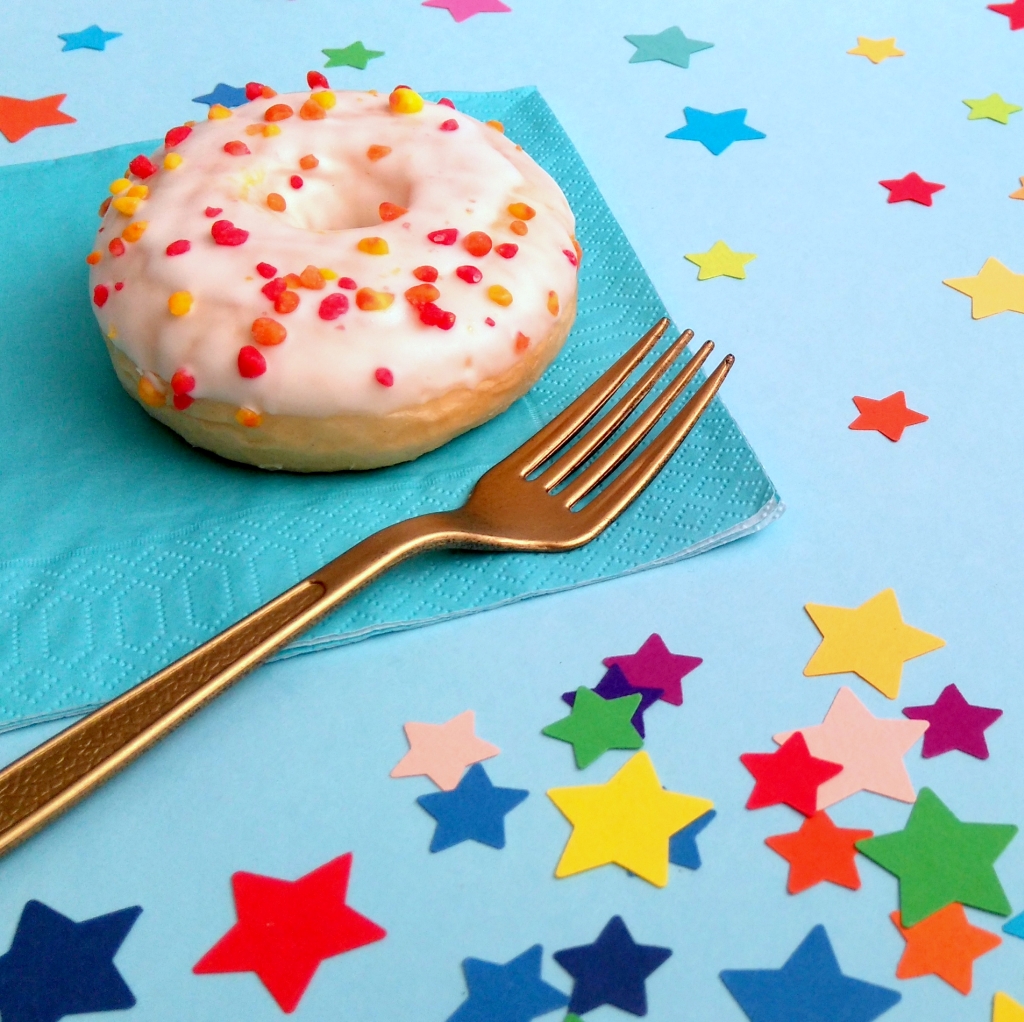 Star confetti is sprinkled on party table with blue tablecloth. A sprinkle iced doughnut and gold fork sit on the table with a blue paper napkin.