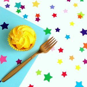 Overhead view of rainbow confetti sprinkled evenly across a party table. An iced cupcake and gold fork are also on the table.