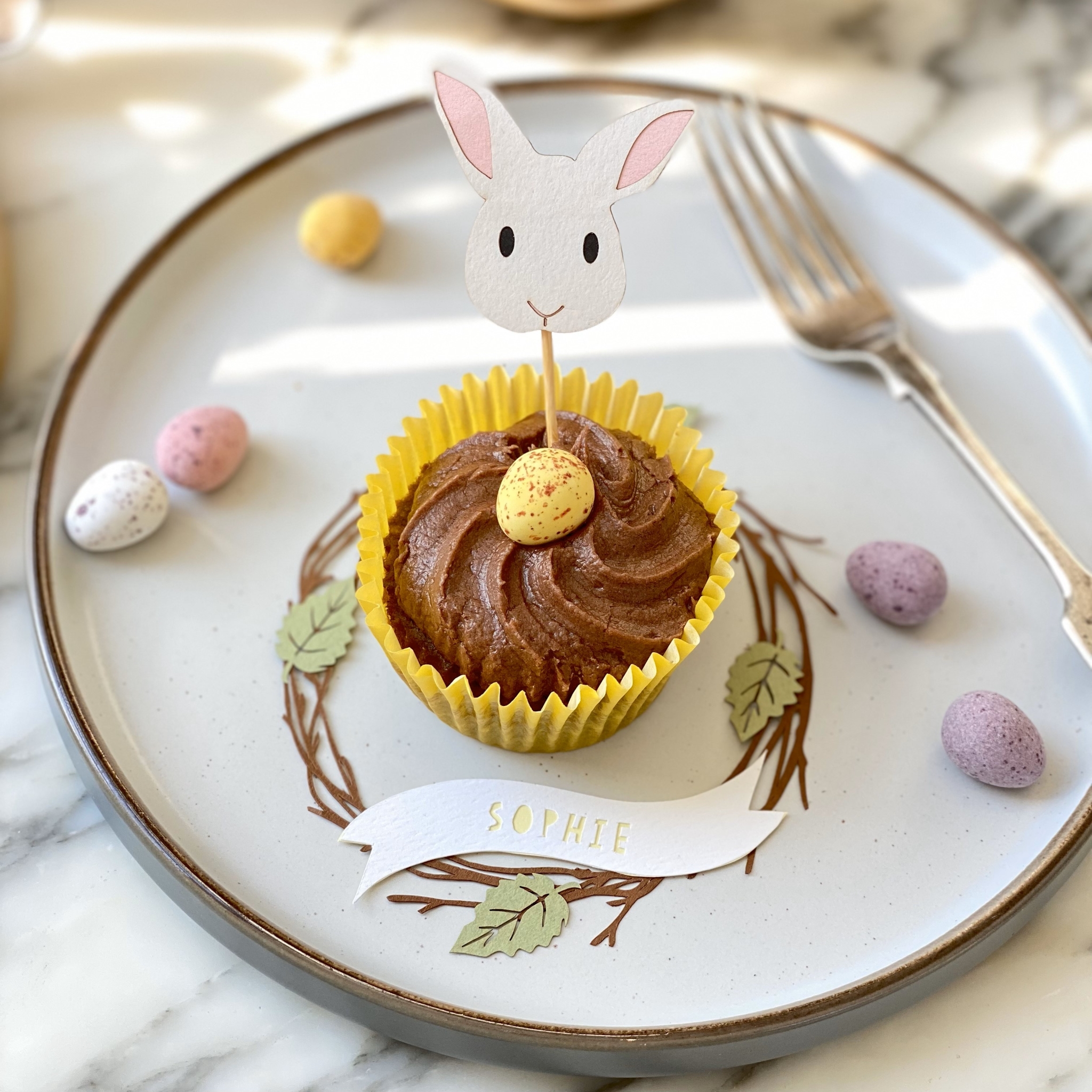 A cupcake topper in the shape of a white bunny face. The bunny has black eyes and pale pink ears. The decoration is placed in the centre of a chocolate cupcake resting on a white plate. Surrounding the cupcake is a papercut birch wreath place setting personalised with the name Sophie.