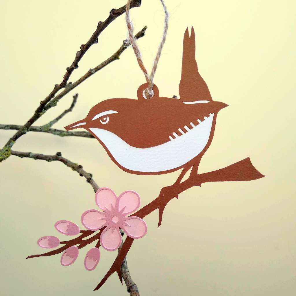 Brown papercut wren with white eye, eye stripe, belly and tail stripe sits on brown branch decorated with pink blossom