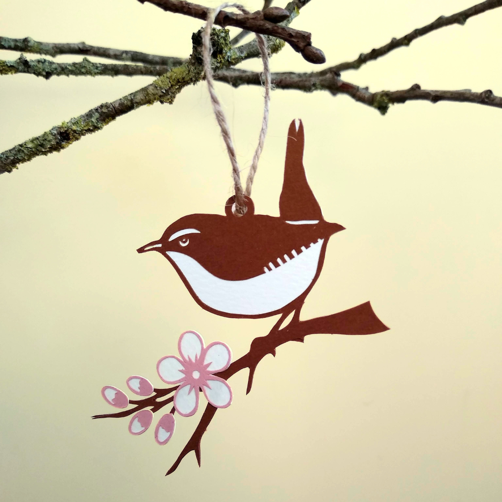 Brown papercut wren with white eye, eye stripe, belly and tail stripe sits on brown branch decorated with white and pink blossom