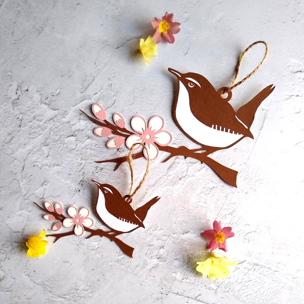 Large and small wren papercut hanging decorations. Brown and white wrens sit on brown branch with blossom. Blossom is white with pink edges.
