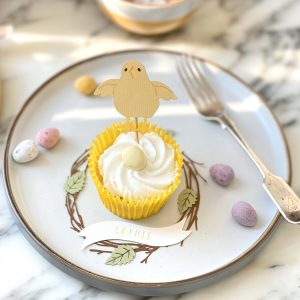 Pale Yellow Easter Chick Cupcake Topper placed into a cupcake decorated for easter. The cupcake is sitting inside a papercut birch wreath place setting decoration displaying the name Sophie. Both decorations sit on a plain white plate along with mini egggs and a fork.