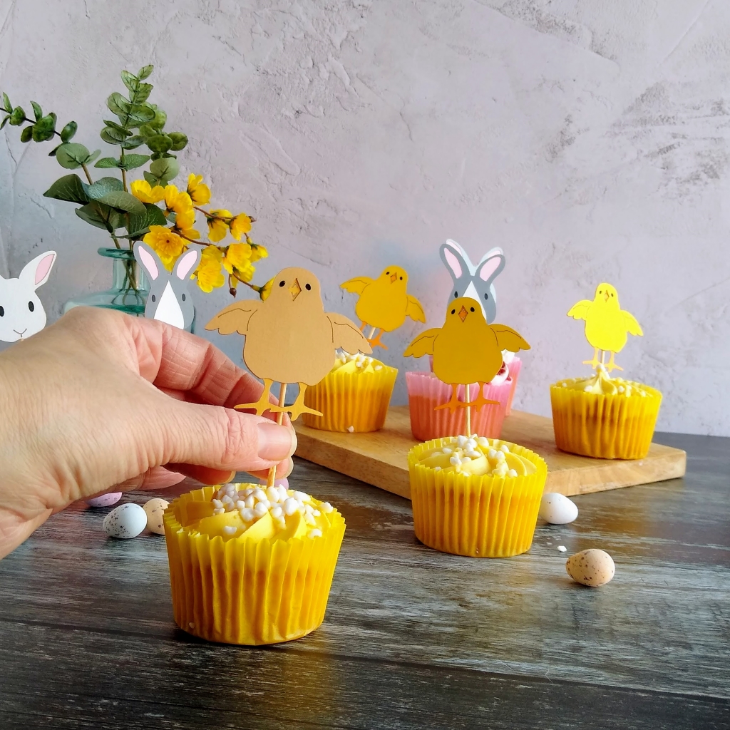 Model places a yellow paper chick cupcake topper into a cupcake. Displayed behind this cupcake are a selection of cupcakes with chick or easter bunny decorative toppers.