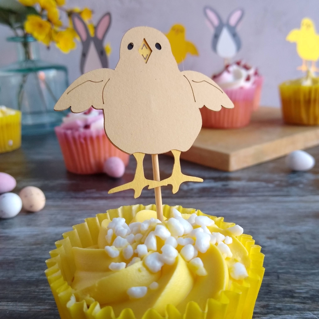 A pale yellow paper cupcake topper shown closeup inserted into a yellow cupcake. The chick has small black eyes, a bright yellow open beak and bright yellow legs.