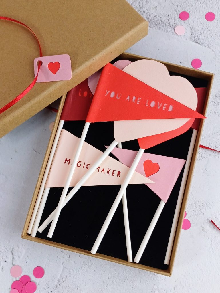 A small box lays open with it's lid to one side. In the box are pink and red paper flags with papercut messages "you are loved" "magic maker" as well as heart shapes 