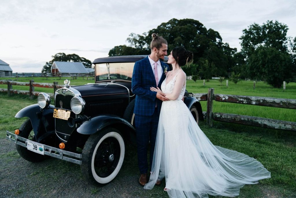 Bride and Groom stand next to black vintage car - license plate"Bumble" in front.