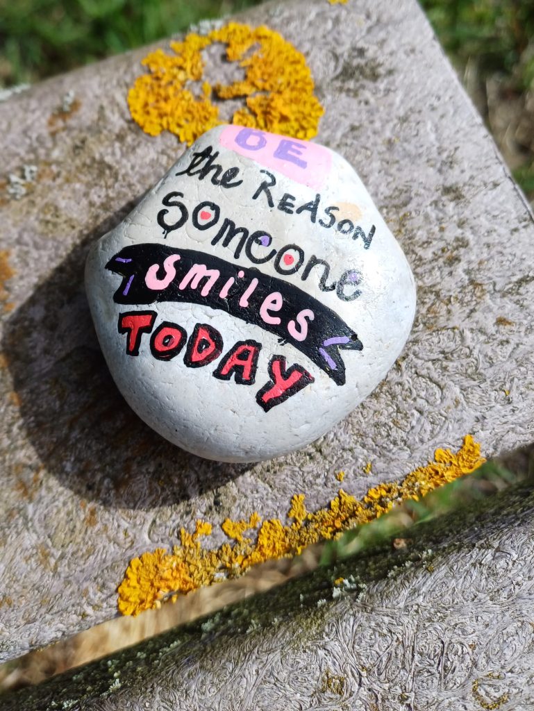 A white rock resting on a wall that has been hand painted with the message "be the reason someone smiles today" in various decorative fonts
