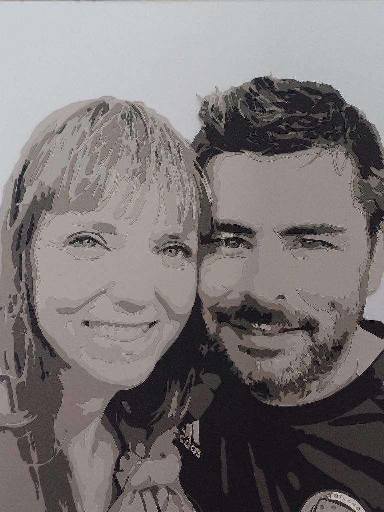 A monochrome papercut portait of a woman and her husband (head and shoulders) stood together smiling at the camera.