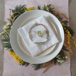 Lasercut birch twig wreath made from dark brown art paper, with 4 pale green paper leaves distributed evenly around the wreath. At the bottom of the wreath is a white paper banner with a name Sophie cut out to reveal the letters in yellow. Place settings is placed on a napkin, on top of stacked plates which sit on a circle of spring flowers and foliage.